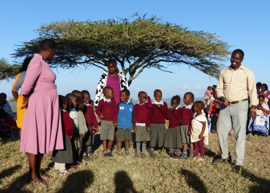 Dunkelhäutige Jungen und Mädchen stehen mit Erwachsenen vor einem Baum mit breiter, flacher Krone und einer weiteren Gruppe von Erwachsenen und Kindern.