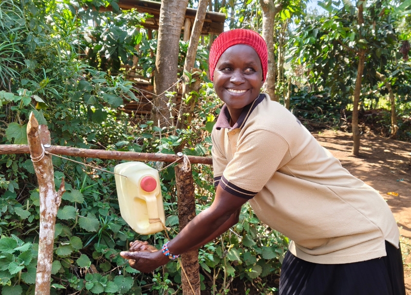 Eine Frau hälte ihre Hände unter einen Kanister, der in ein Holzgestell vor einem Busch eingespannt ist.