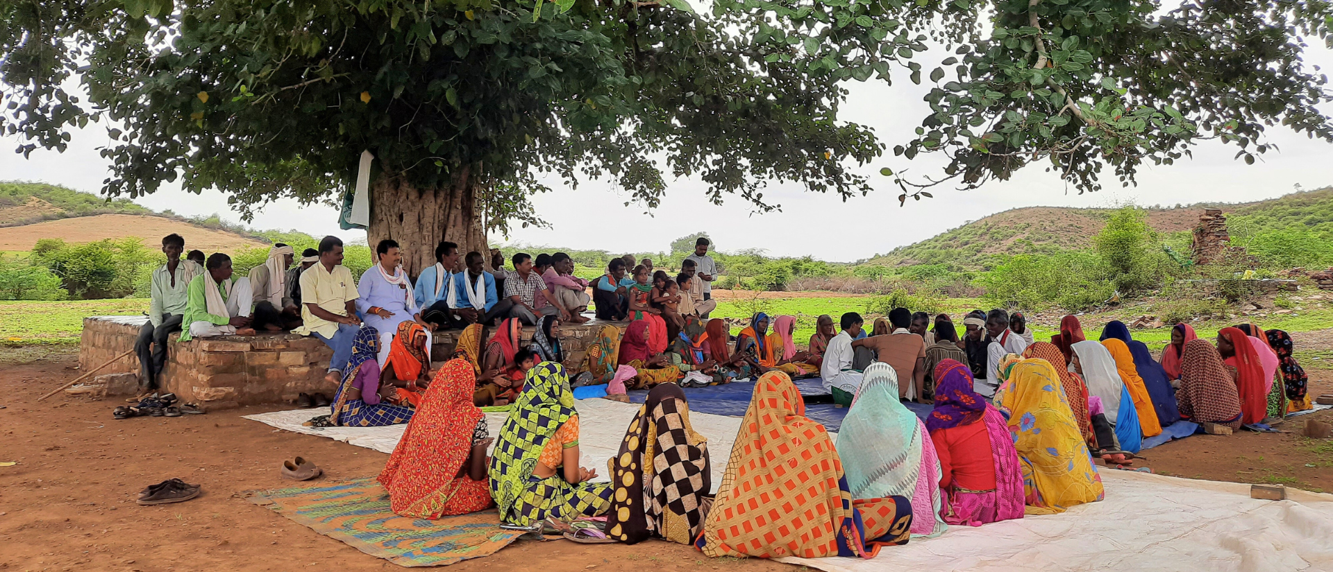 Eine große Gruppe von Menschen hat sich im Schatten eines weit ausladenden Baumes in einer ländlichen Landschaft in Indien versammelt. Einige sitzen auf einer niedrigen Mauer unter dem Baum, andere auf großen Planen und Decken, die auf dem Boden ausgebreitet sind.