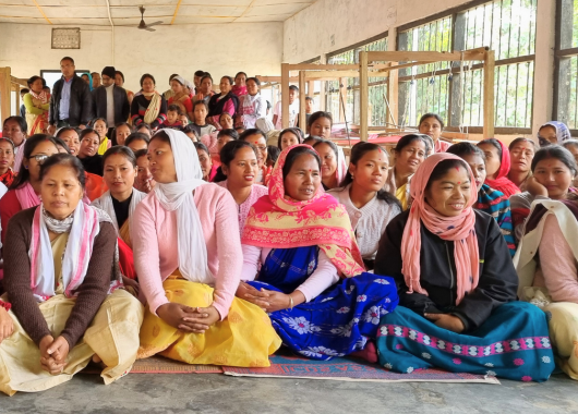 Viele Menschen füllen den Raum. Am rechten Bildrand stehen am Fenster mehre große Webstühle. 