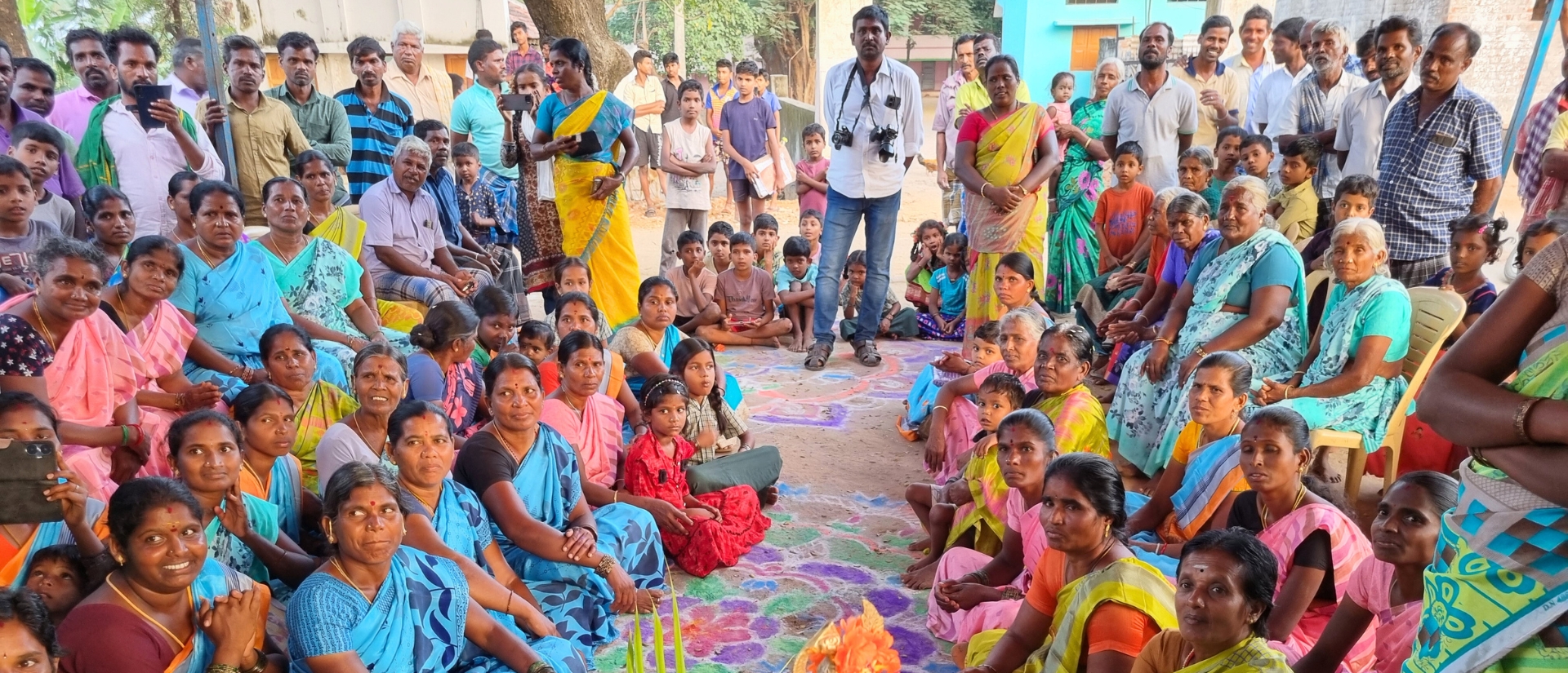 Eine Gruppe von Frauen in bunten Gewändern sitzen in einem Halbkreis auf mit Ornamenten verziertem Sand, im Hintergrund Männer und Kinder