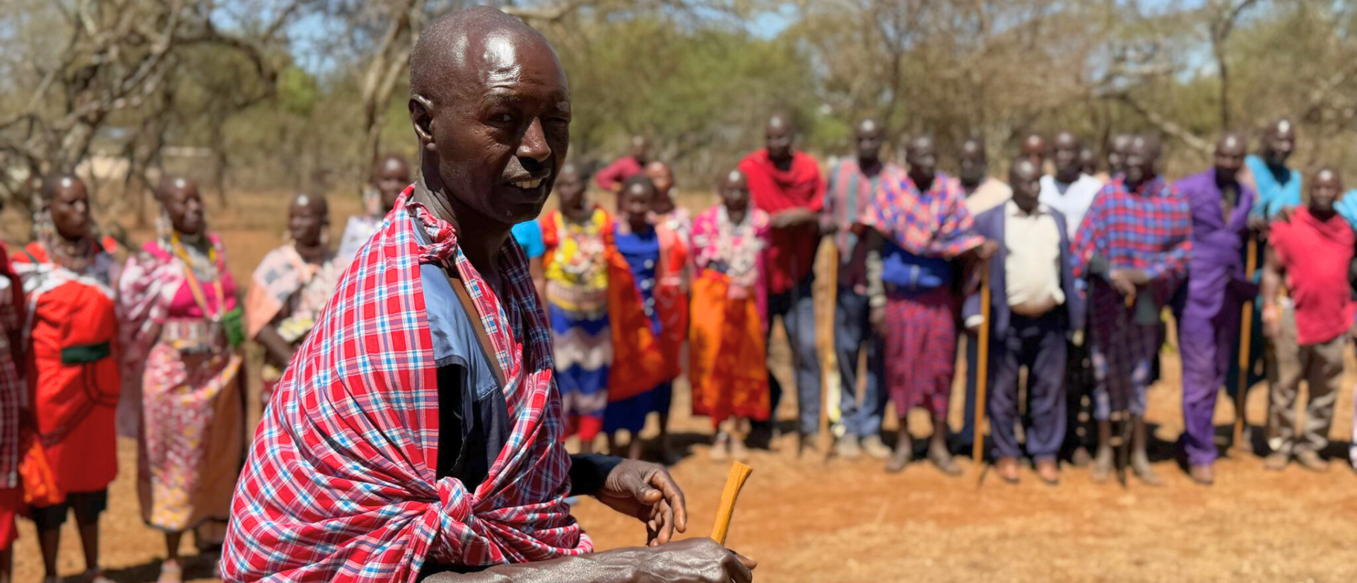 Gruppe von Erwachsenen im Freien vor einer Buschlandschaft mit roter Erde, trockenen Sträuchern und Bäumen. Einige Personen tragen bunte, traditionelle Tücher, andere westlich geprägte Kleidung. Im Vordergrund steht eine Person mit rot-blau kariertem Tuch und einem Stock in der Hand.