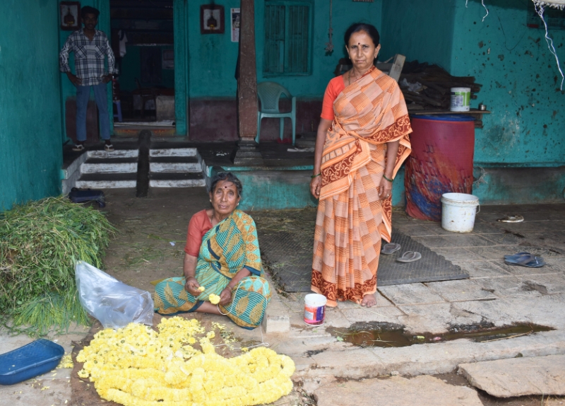Eine Frau sitzt auf dem Boden vor gelben Blüten, die sie auffädelt, neben ihr steht eine weitere Frau, im Hintergrund ein Mann.