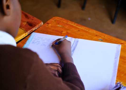 Eine Person sitzt an einem Tisch und schreibt mit einem Kugelschreiber in ein aufgeschlagenes Heft.