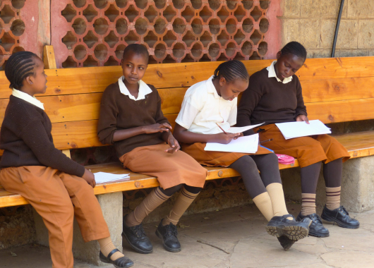 Vier Kinder sitzen draußen auf einer Bank. Zwei haben Hefte auf dem Schoß und einen Stift in der Hand, die anderen beiden haben eine Heft zwischen ihnen liegen.