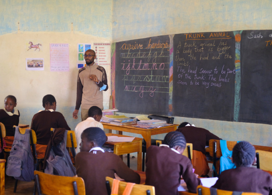Vorderer Bereich eines Klassenzimmers mit Tafeln voller auffälliger, bunter Kreideschrift. Davor steht ein Mann mit Kreide in der Hand neben einem Tisch auf dem viele bunte Hefte liegen. In mehreren Reihen sitzen Schüler*innen an Tischen und Stühlen.