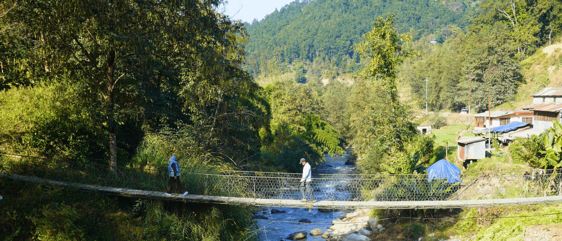 Zwei Menschen kommen sich auf einer Hängebrücke über einem Fluss in einer hügeligen, grünen Landschaft entgegen.