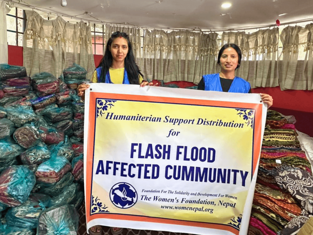 Zwei junge Frauen halten ein Plakat mit dem Titel "Flash Flood Affected Community", im Hintergrund sind Stapel mit Decken und eingetüteter Kleidung zu sehen.