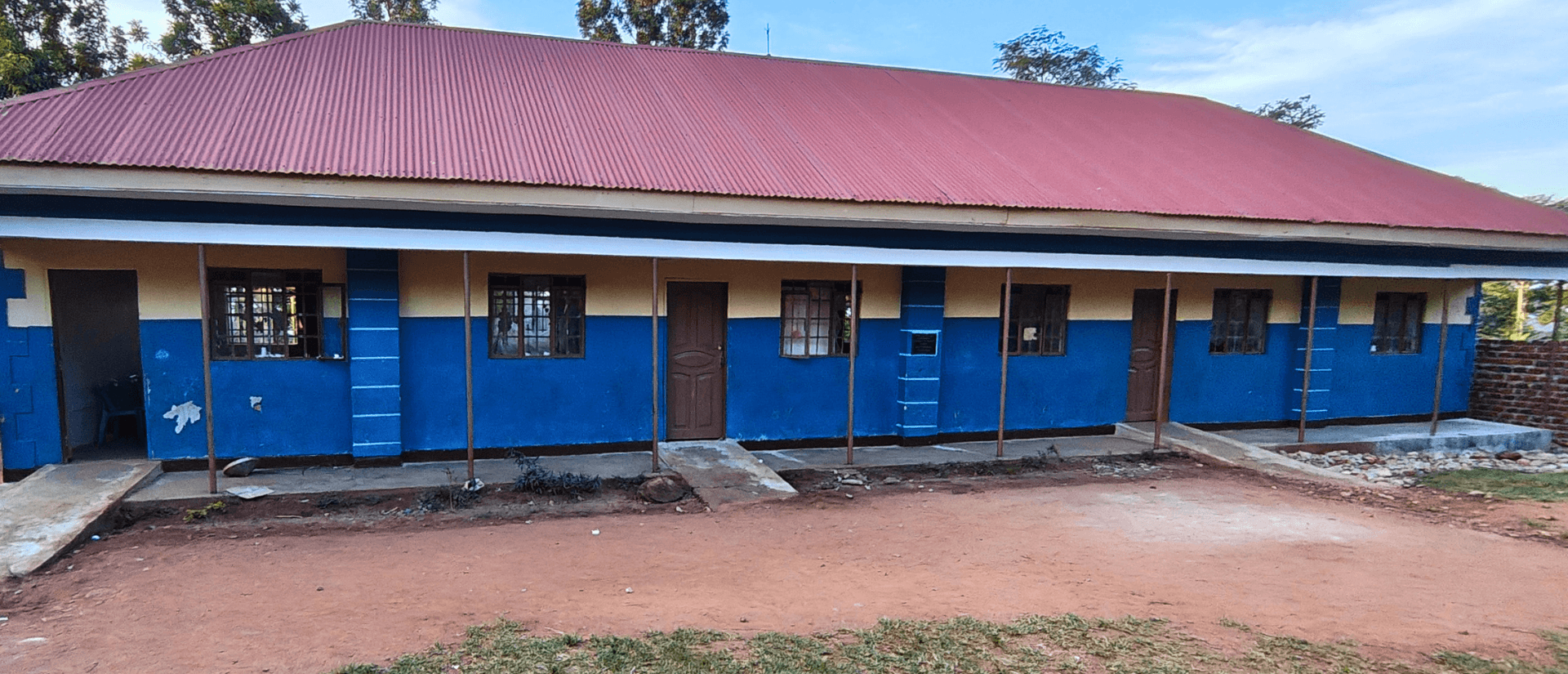 Auf einem Platz mit roter Erde und Gras steht ein leeres Gebäude einer Schule in Uganda.