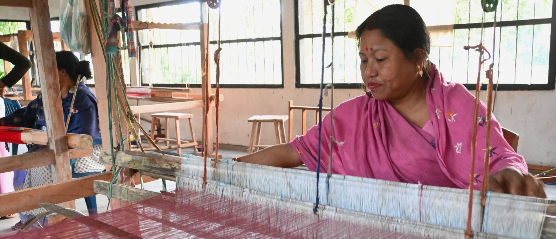 Eine Frau in rosafarbenen Gewand bedient einen Webstuhl, über den weißes und rotes Garn zusammenläuft.