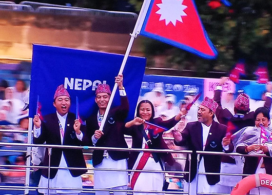 Vier Männer und eine Frau in festlicher nepalesischer Kleidung stehen unter der nepalesischen Flagge auf einem Schiff, schwenken die Flagge und jubeln in die Kamera. Im Hintergrund auf einem Banner steht "Nepal" geschrieben.