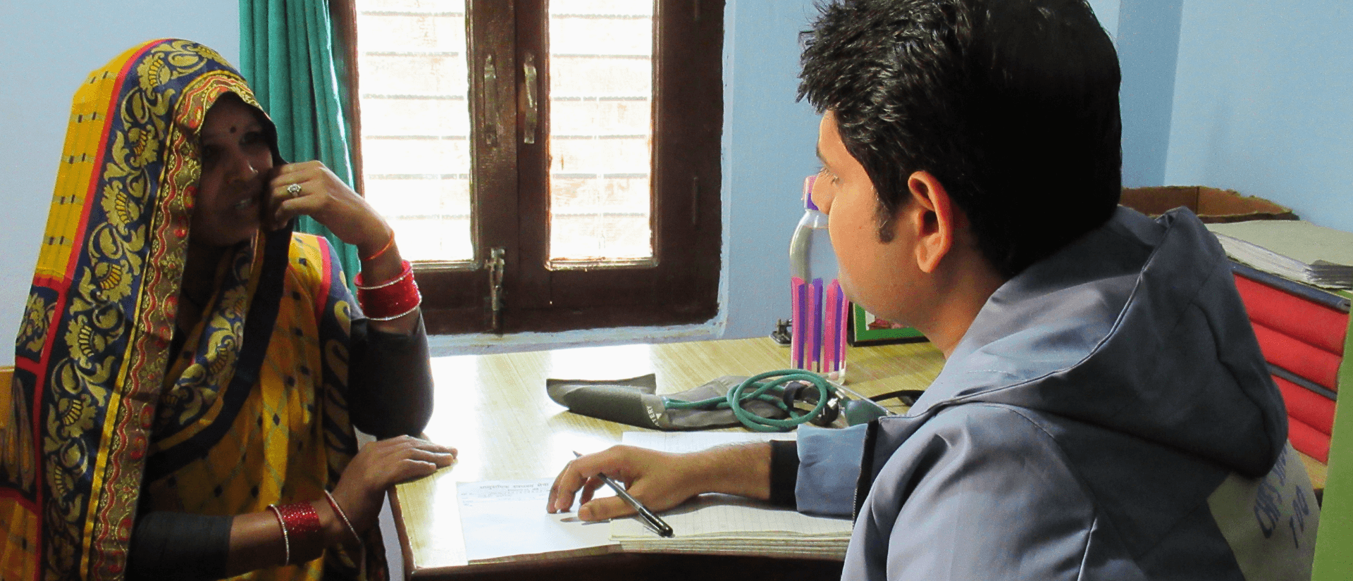 Eine Frau sitzt im Gespräch mit einem Mann an einem Tisch mit Blutdruckmessgerät, der Mann hält einen Stift über einem Notizpapier. 