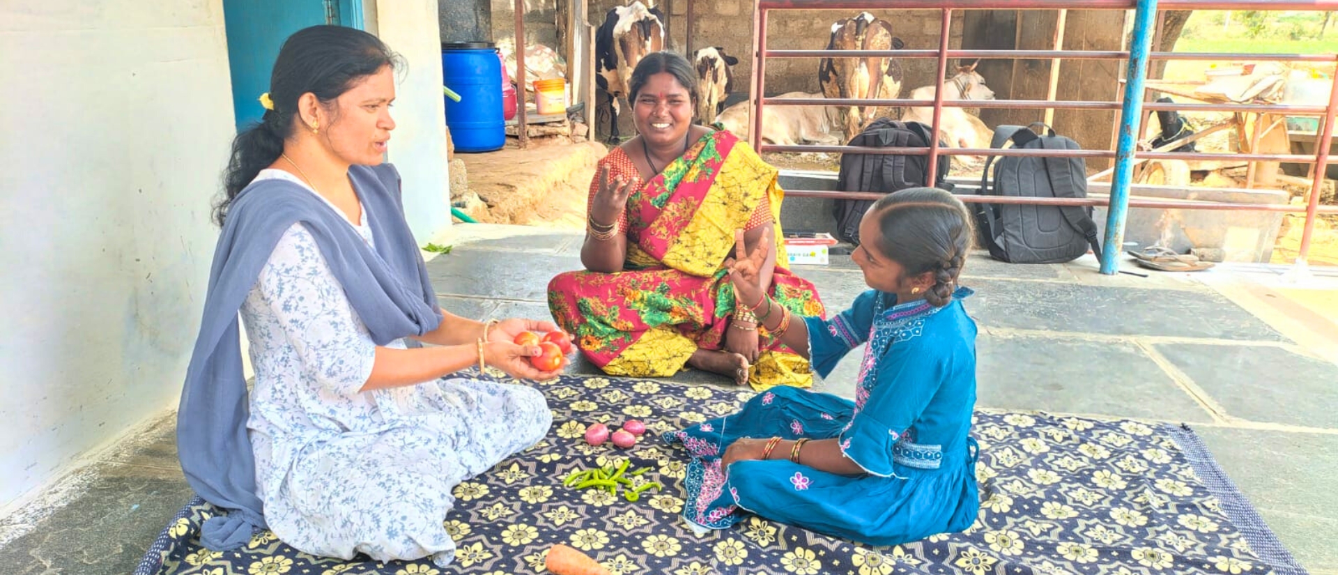 Zwei Frauen und ein Mädchen sitzen im Dreieck, eine der Frauen hält drei Tomaten, das Mädchen und die andere Frau heben drei Finger.