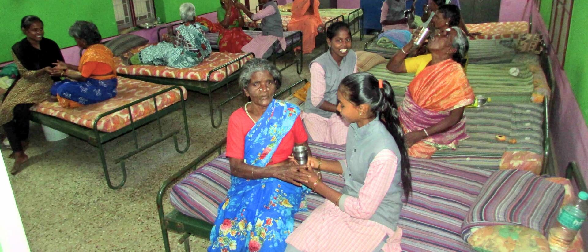 In einem leuchtendgrün gestrichenen Raum sitzen Menschen auf Betten verteilt; im Vordergrund sitzt ein junges Mädchen neben einer Seniorin und hält mit ihr einen silberfarbenen Becher.