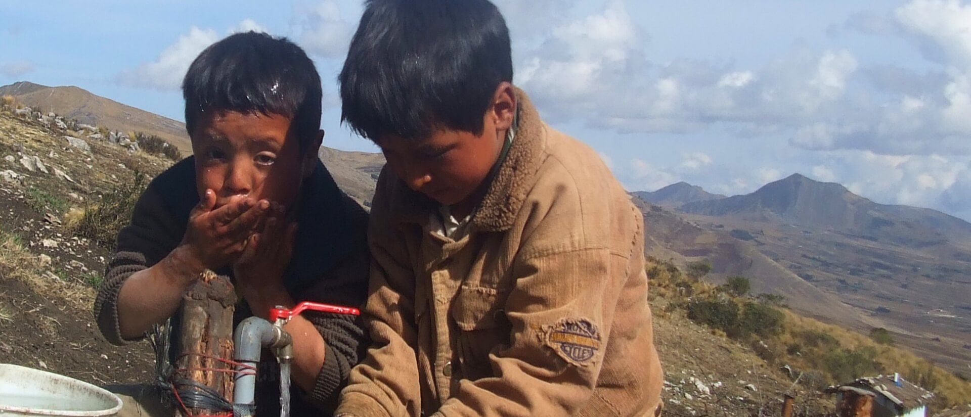 Zwei Jungen stehen an einem Wasserhahn im Freien in einer Berglandschaft, einer trinkt aus der Hand, der andere hält die Hände unter den Wasserstrahl.