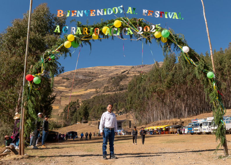 Ein Mann steht vor einer Berglandschaft unter einem großen Bogen aus Zweigen und einem Schriftzug, das zum Agrar-Festival von ACICA willkommen heißt.