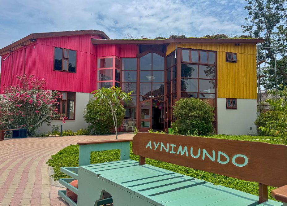 Ein türkisbraune Bank mit dem Schriftzug Aynimundo steht vor einem in Pink, Gelb und Weiß gestrichenen Haus mit großen, in Holz gerahmten Fensterflächen.