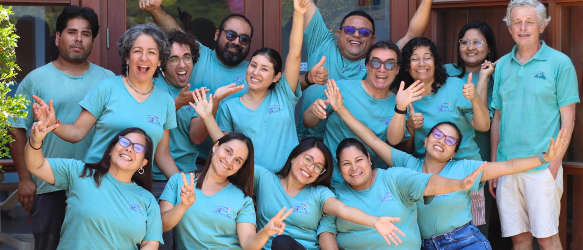 Eine Gruppe von Menschen in türkisblauen T-Shirts lächeln in die Kamera, manche machen mit ihren Armen und Händen Gesten von Jubel und Freude.