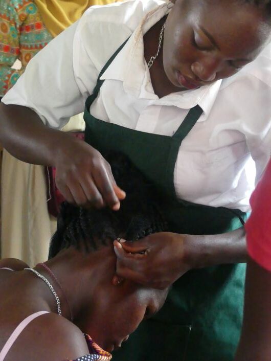 Links unten im Bild sieht man eine Frau mit dem Kopf nach vorne geneigt. Darüber beugt sich eine Frau in Schürze, die der sitzenden Frau die Haare flechtet.