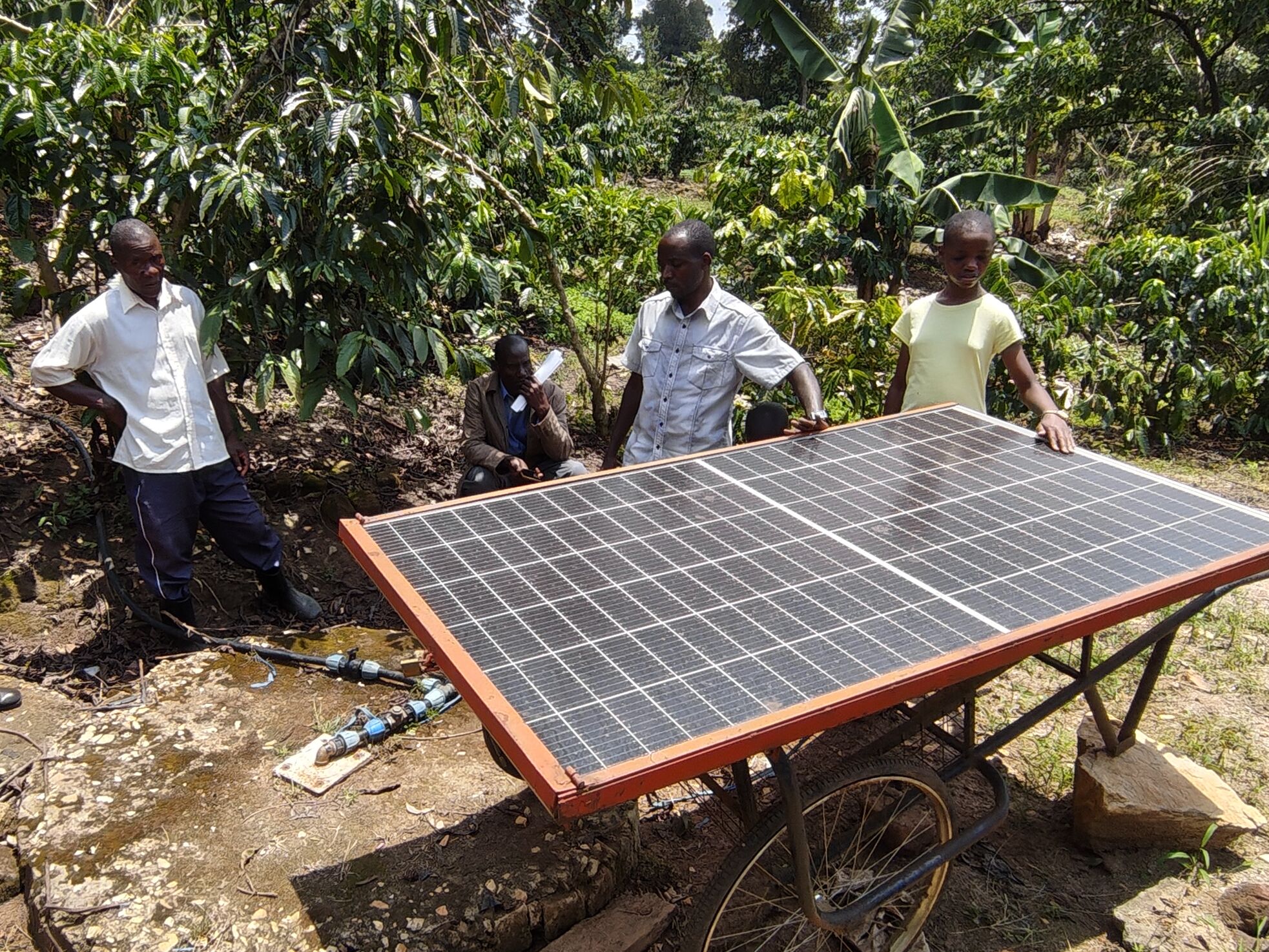 Mehrere Menschen stehen um ein Solarpanel, welches auf einem Gestell mit Rädern liegt. Auf dem Boden führt ein Schlauch in einen runden unterirdischen Wassertank. 