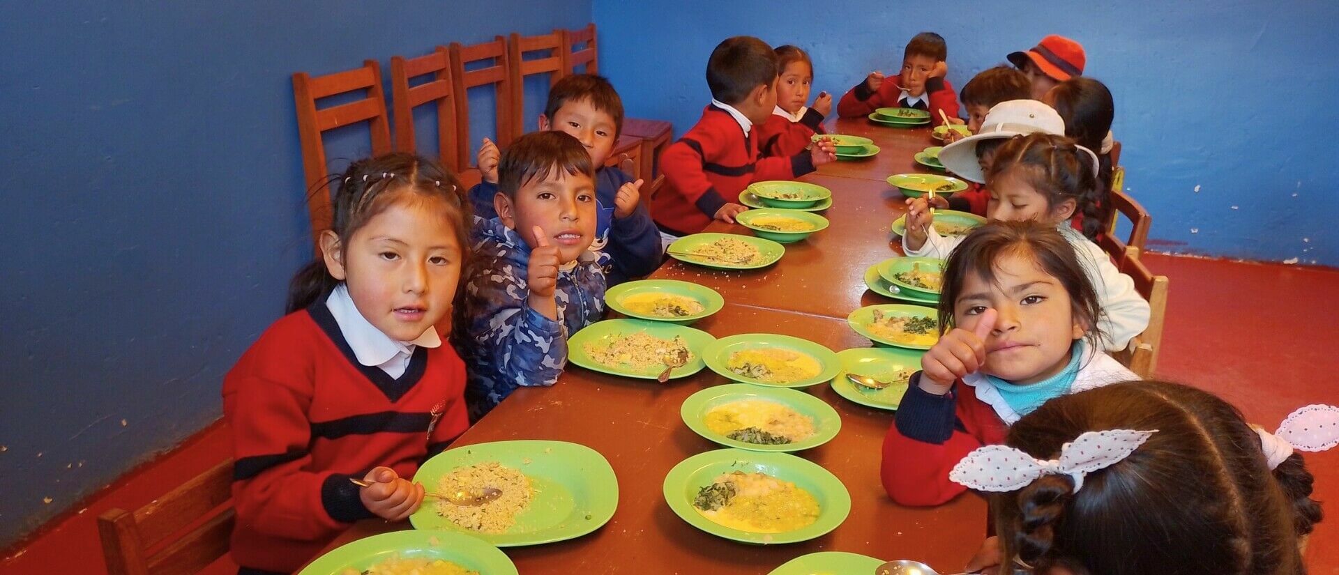Kinder sitzen um einen Tisch vor einer tiefblauen Wand vor ihren Tellern, manche halten einen Löffel in der Hand, andere halten ihren Daumen hoch.