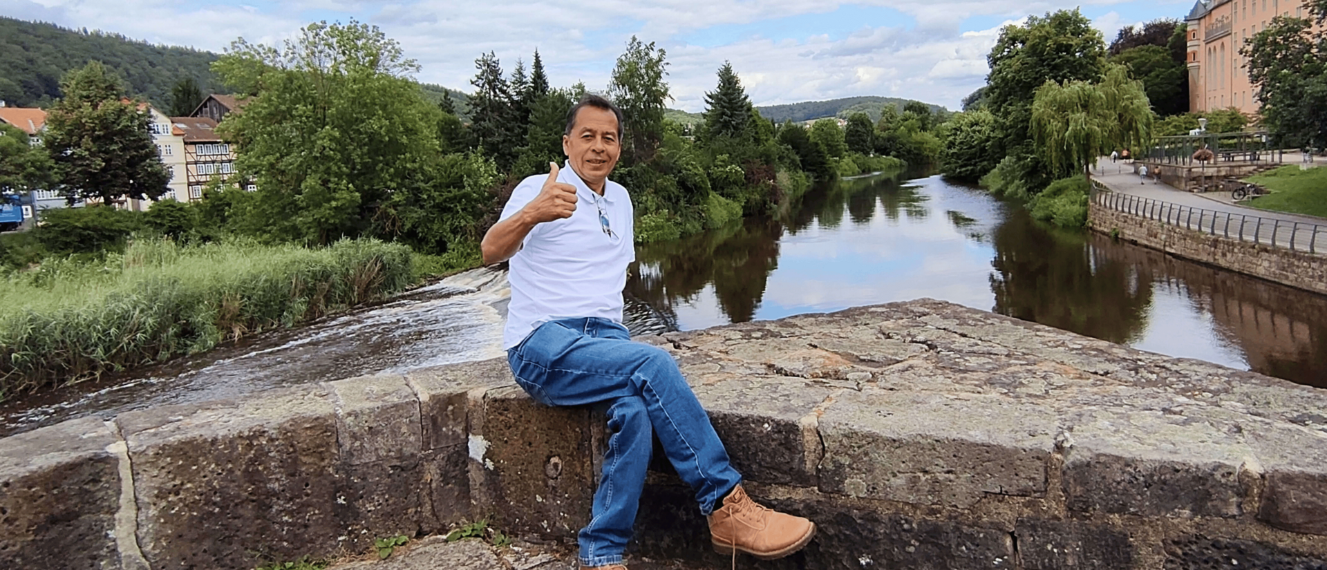 Ein Mann sitzt mit nach oben gestrecktem Daumen auf der Steinmauer einer Brücke über einem Fluss.
