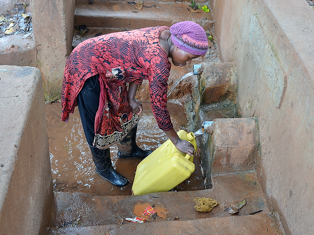Eine Frau aus der Region füllt in gebückter Haltung an der geschützten Quelle Wasser aus Wandrohren in einen gelben Kanister.