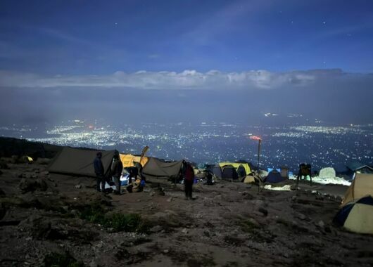 Ein Zeltlager in nächtlichem Licht auf einer Bergebene, dahinter Blick auf ein Tal mit beleuchteten Häusern.