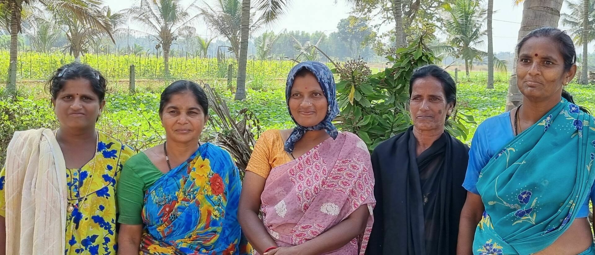 Fünf Frauen stehen lächelnd vor Bäumen und einer mit Pflanzen bewachsenen Fläche.