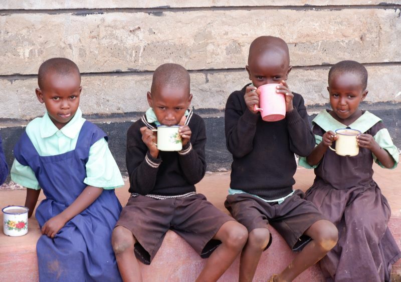 Vier Kinder sitzen nebeneinander. Sie halten in ihren Händen Tassen, aus denen sie zum Teil eine festere Flüssigkeit trinken